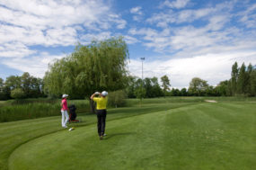 Zwei Menschen spielen Golf bei sonnigem Wetter, wobei einer gerade abgeschlagen hat.