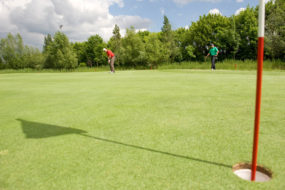 Bild eines Golflochs mit Fahne, im Hintergrund befindet sich ein Mann kurz vor dem Abschlag.