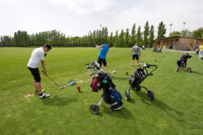 Drei Menschen trainieren mit ihren Ausrüstungen auf der Übungsanlage des Golfplatzes, wobei der erste gerade abgeschlagen hat und der Zweite sich auf den Abschlag vorbereitet.